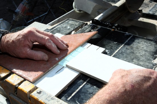 With mosaic tile, a site-built cutting jig can be helpful to support both sides of the cut and hold the tiles stationary. First, cut a saw kerf about two-thirds of the way through a porcelain tile, and use this as a base support. Then use the factory corner (red tile in this photo) to hold the mosaics parallel to the blade at the desired width, and hold the mosaics stationary with a third tile.