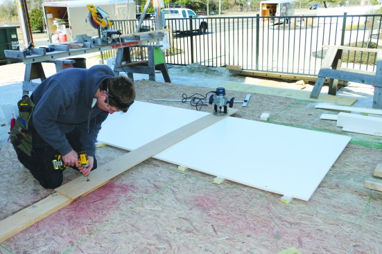 The cap for the curved panel walls is cut from 3/4-inch-thick PVC sheet stock. With the router mounted on the same trammel arm that he used before, a crew member first sets the radius for the outside of the curved cap rail and drives a screw to secure the pivot point.