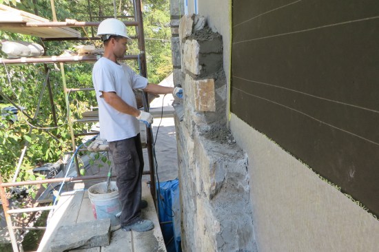 After applying the self-adhering housewrap and 30-lb. asphalt felt paper, a drainage matrix (with gray-colored filter fabric) is installed. Here, Obdyke Slicker Max drainage matrix was used, though on similar masonry veneer projects, the author also uses Mortairvent. The stone veneer is re-installed, secured with stainless steel masonry anchors.
