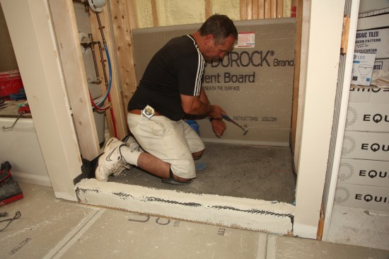 Many tile installers put the backerboard on before installing the mud base, which is acceptable if the board is kept off the surface of the pan—where it could wick up moisture and cause discoloration of the tiles. But this method requires the installer to step on the membrane, potentially exposing 
it to damage. 
Instead, wait until after the mud base is installed to install the backerboard, so that you never have to step on the membrane. With this method, the board can sit directly on top of the mud layer. Attach the board with 1 1/2 -inch galvanized roofing nails, keeping all the nails at least 2 inches above the top of the threshold.