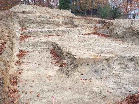 The bottom of the foundation excavation is stepped, with the rear section of the house to be built over a crawlspace to avoid needing to remove additional soil from the lot. The small amount of usable excavated soil can be seen piled behind the foundation hole.