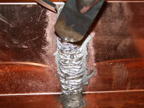 …hold the iron at a shallow angle with the tip against the metal, press the solder stick against one facet of the tip until a drop of melted solder forms, then use the iron to spread the drop across the seam. Each drop adds another layer, and the heat of the iron on the copper draws the solder into the seam to make a water-tight seal. The thick “ribs” reinforce and strengthen the seam. Move up a little bit and repeat; building up the joint as you go along.