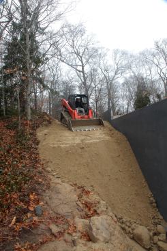 Using his skid-steer machine, George gradually continues to fill beside the foundation, again using the bucket to slope the fill away from the foundation and to follow the existing land contour.