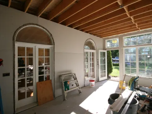 After the rough wiring was completed in the sunroom, we insulated the knee walls, the open space in the posts, and the stud bays of the gable-end wall of the existing house with “Thermoseal” open-cell spray foam (hitting the gaps at the windows with low-expanding foam). We sprayed the whole roof assembly as well, leaving the sunroom’s ceiling uninsulated. After hanging &frac12;-inch drywall on the posts and all of the walls, we converted the two exterior doors in the existing gable-end wall into interior doors. This involved removing existing weatherstripping and the exterior sills