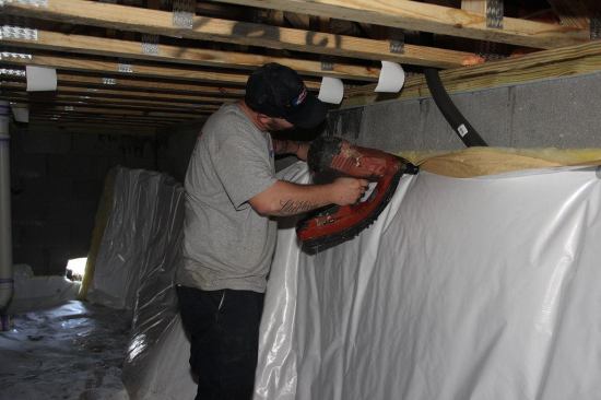 A crew member fastens the fiberglass insulation blanket to the block wall using gun-driven masonry nails with metal disk washers. The space between the top of the insulation blanket and the framing above is required by code, to allow inspection for termite tubes. Termites are a significant risk in coastal South Carolina.