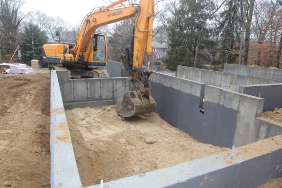 After dumping clean fill inside the garage foundation with the excavator, George then uses the bucket to compact the fill. He also uses a hand-operated compactor on the soil.