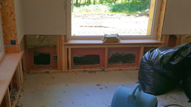 Here is a view of the wall during remediation work after the flood. Drywall has been removed to a point above the water damage line, Roxul insulation has been pulled out of the inboard wall stud cavity, and sections have been cut out of the OSB air barrier in each stud bay to allow access to the outer wood I-joist insulated bays. Note the red color of the fluid-applied air barrier coating applied to the inside of the OSB sheathing during construction.