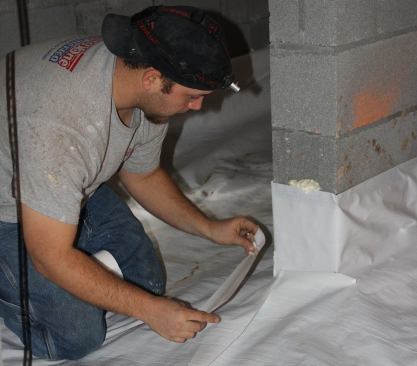 Seams in the poly ground cover are sealed with tenacious vapor-barrier tape. Gun foam fills the gap between the poly and the concrete block in this picture; for air-tightness and vapor control, the crew installs a bead of the expanding adhesive foam at the top seam where the poly meets the block wall around the entire perimeter of the crawlspace.
