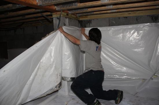 A crew member installs a poly-faced fiberglass insulation blanket around the block wall perimeter of the crawlspace.
