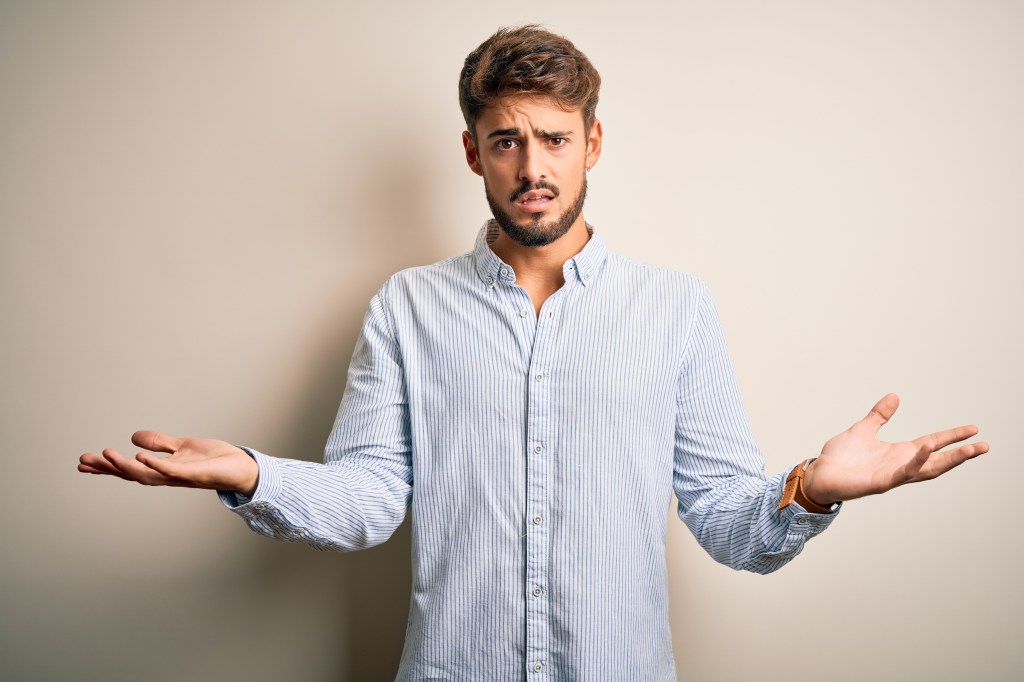 Young handsome man with beard wearing striped shirt standing over white background clueless and confused with open arms, no idea concept.