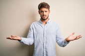 Young handsome man with beard wearing striped shirt standing over white background clueless and confused with open arms, no idea concept.